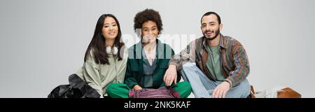 felice multiculturale studenti in abiti alla moda seduta e guardare la macchina fotografica isolato su grigio, banner, immagine stock Foto Stock