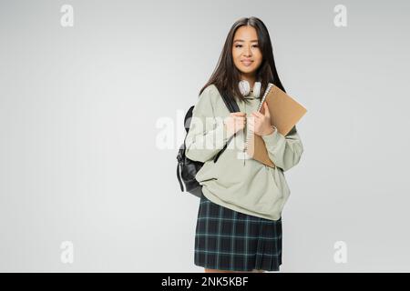 grazioso studente asiatico in felpa con cappuccio e plaid gonna in piedi con copybook e zaino isolato su grigio, immagine stock Foto Stock