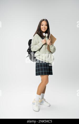 intera lunghezza di donna asiatica felice in elegante vestito in piedi con zaino e notebook su sfondo grigio, immagine stock Foto Stock