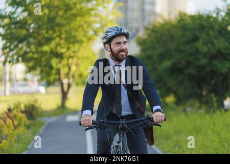Giovane manager maschile in tuta d'affari che si trova al lavoro sulla pista ciclabile di mattina presto. Ritratto di felice bearded executive guidando bicicletta, mentre lookin Foto Stock