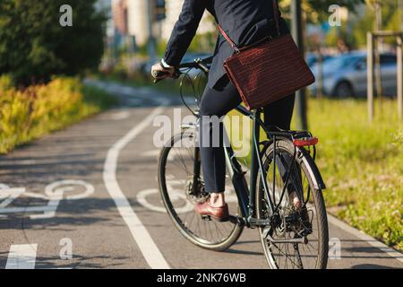 Anonimo maschio manager in business outfit equitazione sulla pista ciclabile in mattinata soleggiata. Vista rifilata di executive con valigetta in pelle per lavorare in bicicletta Foto Stock