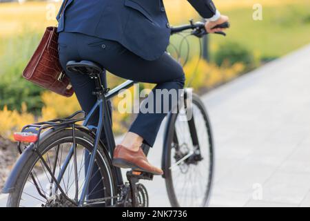 Anonimo imprenditore maschile in bicicletta vestito nero su strada asfaltata di giorno. Vista di raccolto di uomo d'affari in abbigliamento costoso a cavallo in bicicletta in città ar Foto Stock