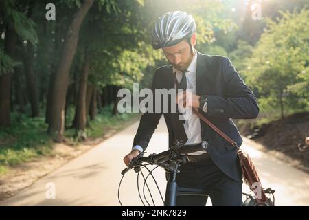 Giovane uomo d'affari in elegante cravatta di fissaggio tuta, mentre si prepara a guidare in bicicletta di giorno. Vista frontale di un uomo serio nel casco di protezione in preparazione Foto Stock