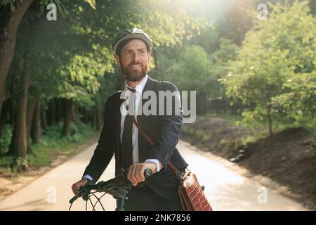 Felice uomo bearded in tuta per lavorare in bicicletta in mattinata di sole. Vista frontale del boss maschile dai capelli scuri in elegante vestito in piedi con la bicicletta, mentre looki Foto Stock