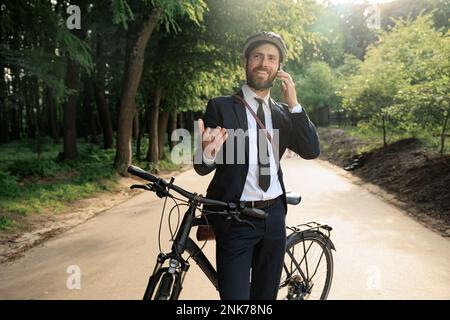 Uomo d'affari di successo che si appoggia sulla bicicletta, mentre si effettua una telefonata sulla strada. Vista frontale dell'uomo felice in tuta che ha stop to call dal telefono cellulare, mentre Foto Stock