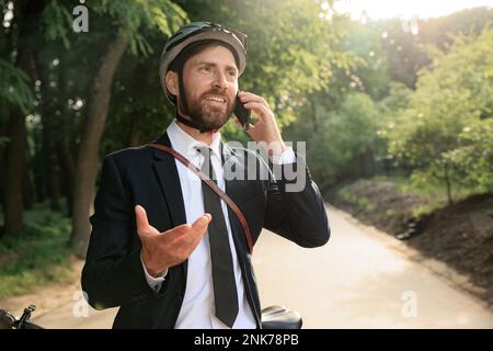 Un dipendente allegro che parla al telefono, mentre si guida in bicicletta per lavorare. Ritratto di felice uomo d'affari brunet in casco protettivo gesturing, durante la realizzazione Foto Stock