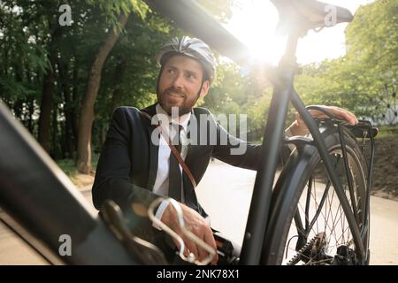 Uomo sorridente in abito elegante che squatting giù per controllare la ruota per bicicletta nel parco cittadino. Ritratto di felice imprenditore maschile in casco che esamina bicicletta, whil Foto Stock