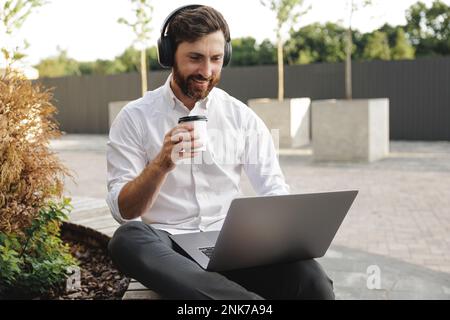 Vista frontale di un bel uomo d'affari in cuffie sedute su panca di legno e lavorando all'aperto. Un maschio sorridente che tiene in mano una tazza di caffè mentre usa por Foto Stock