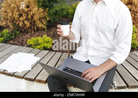 Lavoratore di un ufficio maschile che utilizza un notebook portatile per cercare informazioni mentre si diverte a bere un caffè. Vista ritagliata dell'uomo in abbigliamento formale che lavora all'aperto wh Foto Stock