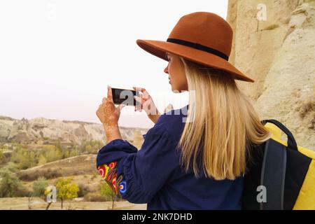 Viaggiare bella donna bionda capelli indossa gonna blu autentica e cappello marrone scattare foto in Love Vally a Goreme, Cappadocia, Turchia. Spazio copia, Foto Stock