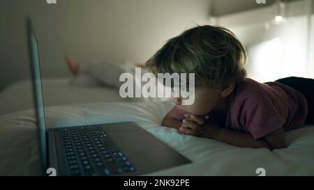 Ragazzino che guarda i cartoni animati che si posano a letto di notte. Bambino ipnotizzato da media online primo piano faccia in serata Foto Stock