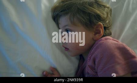 Il ragazzino si stendeva a letto guardando i media di intrattenimento di notte con uno schermo luminoso Foto Stock