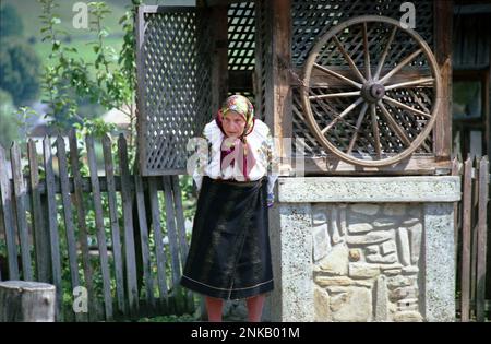 SRAJA, Contea di Suceava, Romania, 1998. Donna anziana che indossa costume tradizionale vicino ad un pozzo d'acqua con ruota. Foto Stock