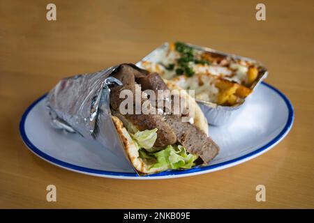 Un giroscopio greco mediterraneo avvolto in un foglio su un piatto con un lato di patatine fritte greche ricoperte di mozzarella e formaggio feta fuso, erbe aromatiche e spezie Foto Stock