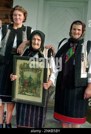 Suceava County, Romania, 1998. Tre generazioni di donne in una famiglia locale in posa nei loro costumi tradizionali. La donna anziana sta tenendo una stampa incorniciata raffigurante il marito come soldato di fanteria nell'esercito austro-ungarico. Foto Stock