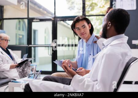 Medico specialista che spiega il trattamento farmacologico al paziente asiatico durante l'esame visita di controllo in ospedale ufficio. Giovane uomo che firma i documenti medici prima di iniziare la consultazione. Servizio sanitario Foto Stock