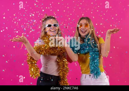 Due giovani donne caucasiche bionde festeggiano, al nightclub con divertenti coriandoli, isolate su sfondo rosa Foto Stock