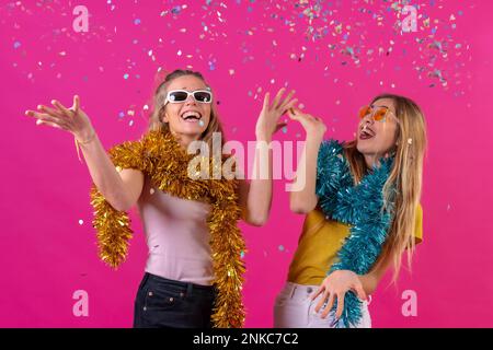 Due giovani donne caucasiche bionde festeggiano, divertendosi a lanciare coriandoli, isolate su sfondo rosa Foto Stock
