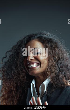 Ritratto di Donna con pelle nera con lunghi capelli ondulati, in elegante camicia bianca e giacca nera, catturata in mezzo a risate o ad un momento intenso Foto Stock