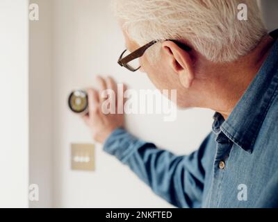 Un uomo anziano che regola il controllo del termostato intelligente sulla parete a casa Foto Stock