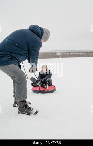 Padre che tira il figlio seduto sulla slitta gonfiabile nella neve Foto Stock