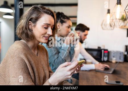 Donna d'affari sorridente che usa il telefono cellulare con i colleghi in background Foto Stock