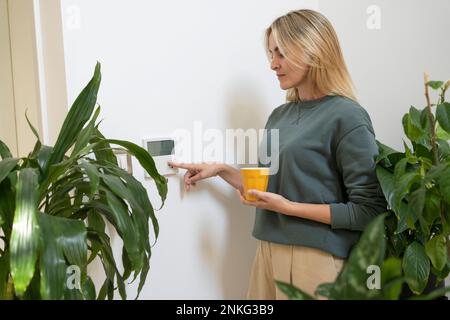 Donna con una tazza di caffè che regola il termostato intelligente a casa Foto Stock