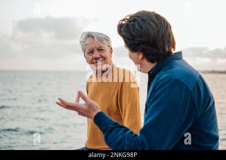 Un uomo anziano sorridente con un figlio che discute di fronte al mare Foto Stock