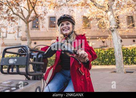 Felice donna matura indossando casco in bicicletta per strada Foto Stock