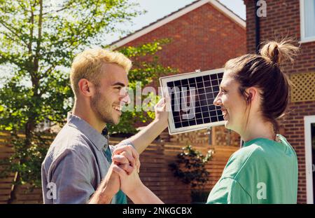 Coppia sorridente con le mani intrecciate tenendo il pannello solare di fronte alla casa Foto Stock