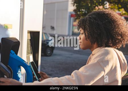 Giovane donna che effettua il pagamento tramite smartphone presso la stazione di ricarica per veicoli elettrici Foto Stock