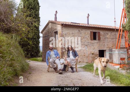 Felice coppia matura seduta su sedia con cane di fronte a casa Foto Stock