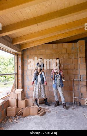 Felice uomo e donna che trasportano le figlie sulle spalle in piedi davanti al muro di mattoni Foto Stock