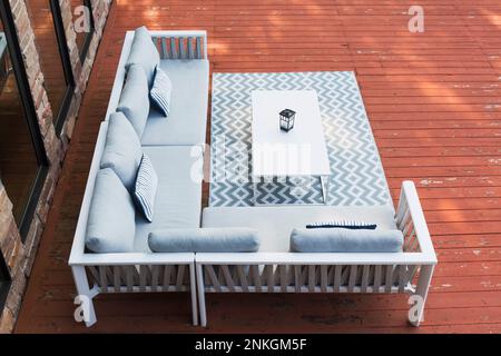 Divani laccati bianchi con cuscini da salotto grigi, cuscini a righe blu e tavolino da caffè sul tappeto sulla terrazza del patio in legno color marrone. Foto Stock