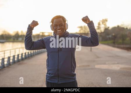 Felice uomo anziano che indossa cuffie che flettono i muscoli sul lungomare Foto Stock