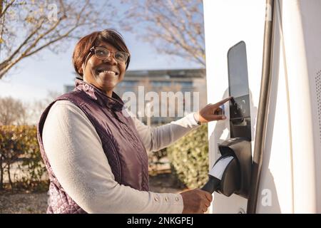 Donna anziana sorridente e premurosa in piedi accanto alla stazione di ricarica elettrica Foto Stock