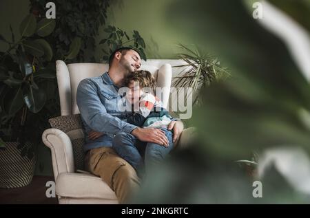 Un padre sorridente che si rilassa con un figlio in poltrona a casa Foto Stock