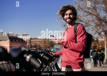 Giovane uomo sorridente con smartphone in piedi presso il sistema di condivisione di biciclette elettriche Foto Stock