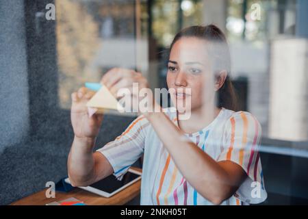 Giovane donna d'affari che appiccicano le note adesive sul vetro insonorizzato in ufficio Foto Stock