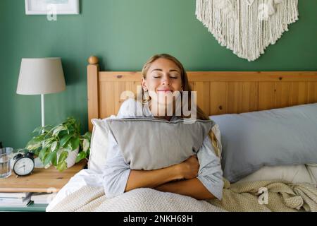 Donna sorridente con gli occhi chiusi che abbraccia il cuscino a letto a casa Foto Stock