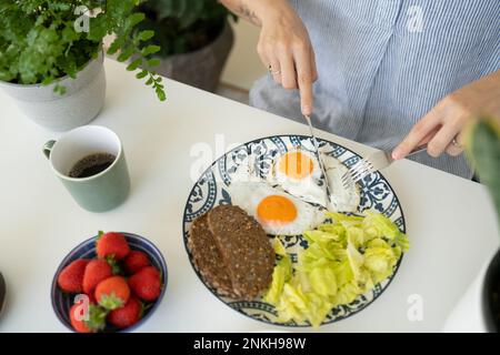 Mani di donna che tagliano l'uovo fritto con il coltello a casa Foto Stock