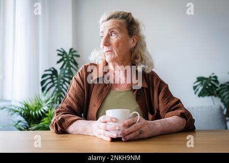 Donna anziana preoccupata che tiene la tazza seduta a casa Foto Stock