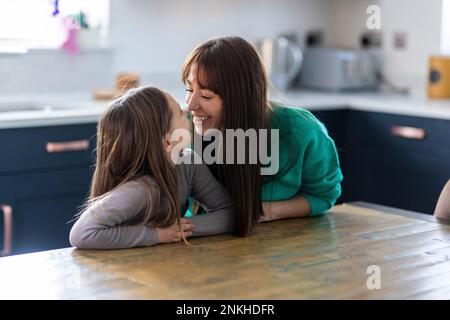 Madre felice abbracciando la figlia seduta al tavolo in casa Foto Stock