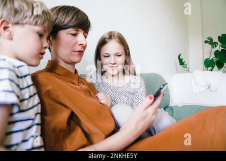 Donna che guarda lo smartphone con un figlio e una figlia seduti a casa Foto Stock