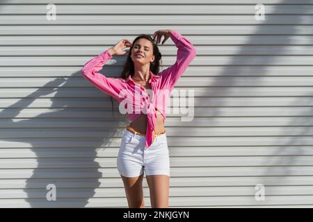 Giovane bella donna brunetta in una camicia rosa e pantaloncini in denim bianco sullo sfondo di un recinto porta garage leggero Foto Stock