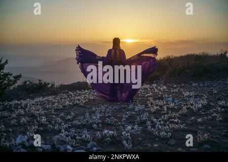 Tramonto viola abito donna montagne. Ascesa del mistico. tramonto sulle nuvole con una ragazza in un lungo vestito viola. Nel prato c'è un prato Foto Stock