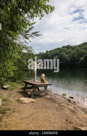 Due bambini sul lago. Una in piedi su una panca da picnic con canna da pesca Foto Stock