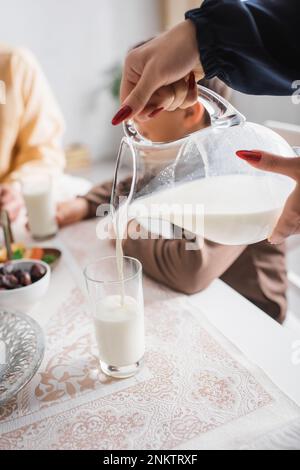 Vista ritagliata di una donna musulmana che versa latte in vetro vicino alla famiglia sfocata al mattino, immagine stock Foto Stock