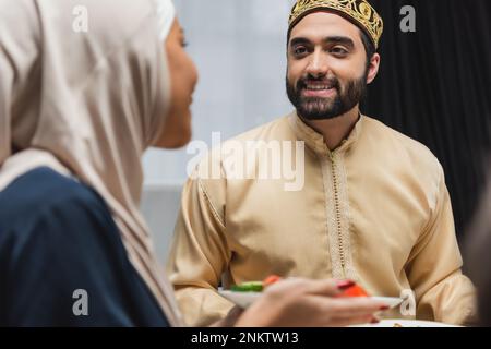 Uomo musulmano positivo che guarda la moglie sfocata con il cibo a casa, immagine di scorta Foto Stock