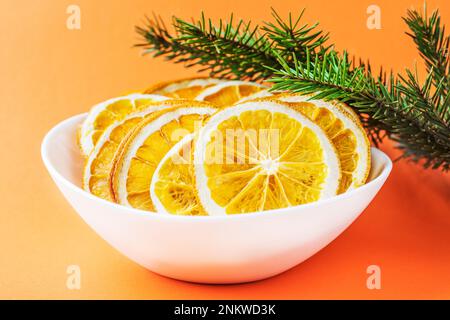 Scaglie di arancio in un piatto bianco, vicino al ramo di abete rosso, su sfondo rosso arancio. Eco biologico fatto in casa fette di arancia secca, patatine fritte. Snack sano. Cl Foto Stock